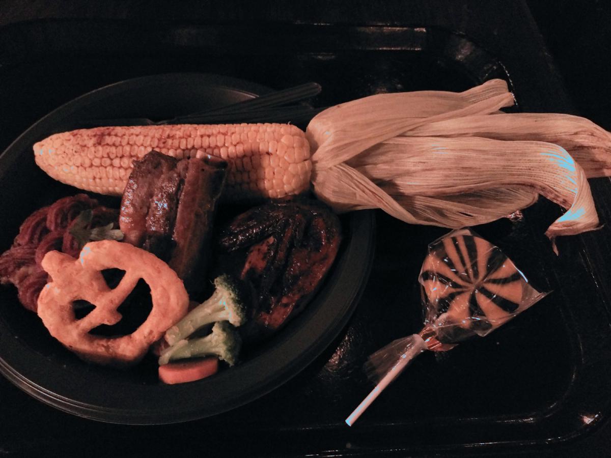 黒いお皿に乗った肉料理、皮付きのコーン、かぼちゃ型のパイ、野菜。右には黒とオレンジの渦巻き模様の棒付きキャンディ。